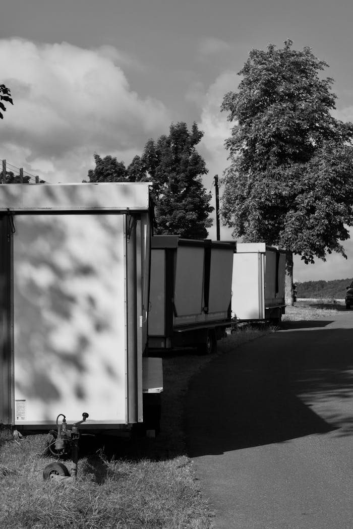 Monochrome image of trailers lined along a rural road under a bright sky.